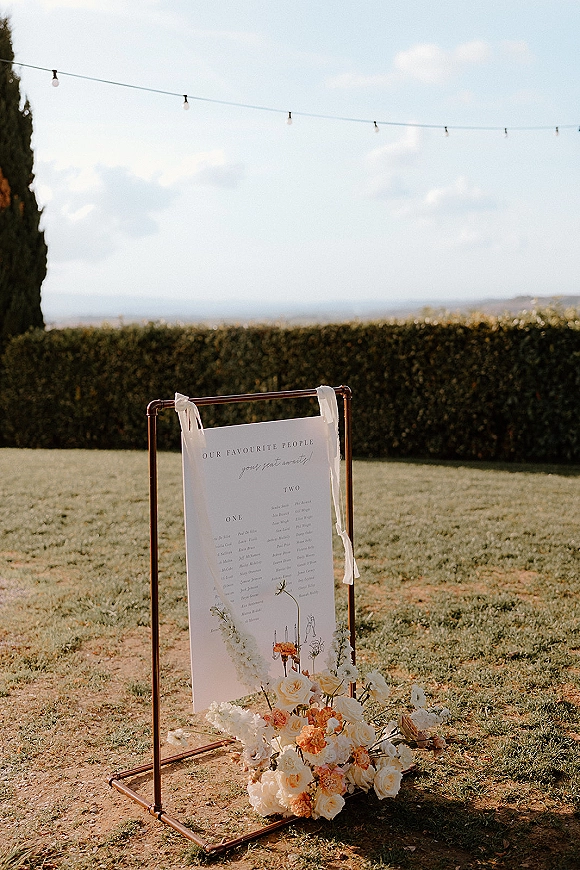 Wedding seating chart seating chart sign on a copper stand with ribbon ties and rose florals beneath, set on a lawn with string lights