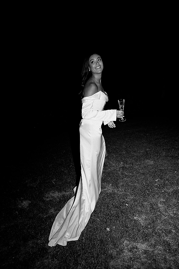 Bridal portrait in a black and white wedding portrait style, bride looking over her shoulder holding a champagne flute on a lawn at night