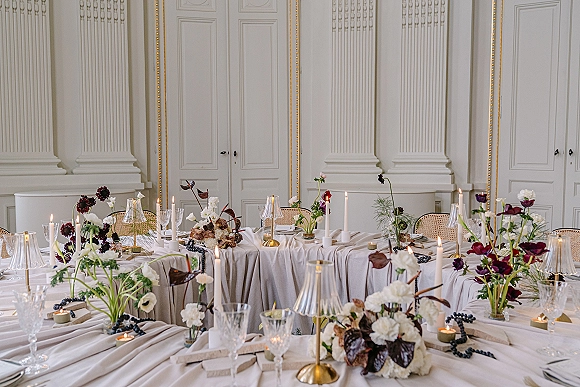 Reception tablescape with wedding head table decor, taper candles, mini table lamps, burgundy florals, grapes, and cane chairs in a white-paneled room