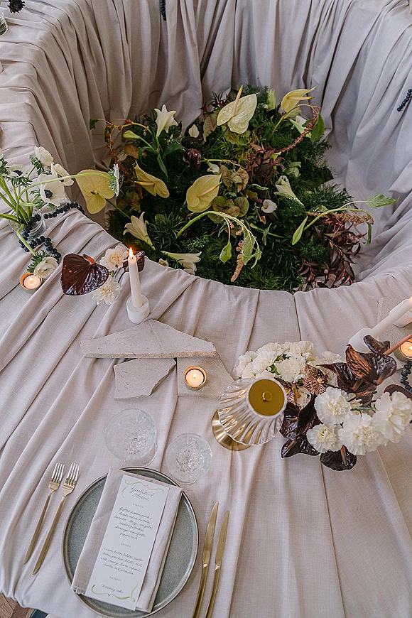 Reception tablescape with a wedding table centerpiece of anthurium and white flowers, taper candles, gold flatware, and draped linen backdrop