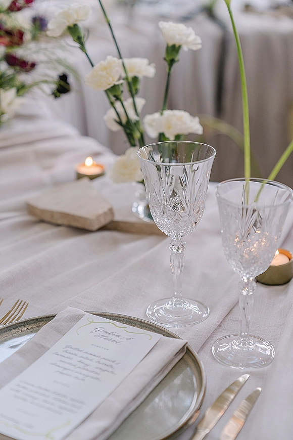Reception tablescape with wedding place setting, menu card on linen napkin, crystal goblets, gold flatware and taper candle against white drapes