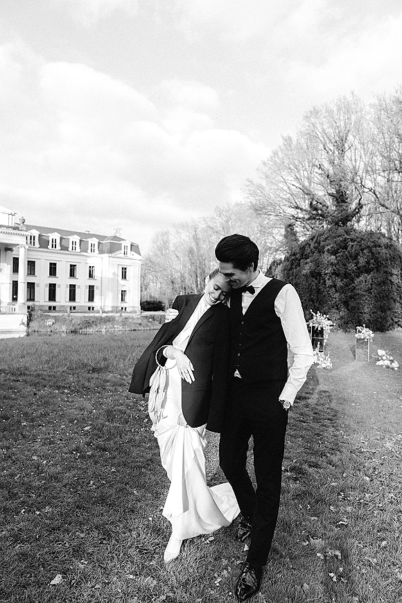 Couple portrait in a black and white wedding portrait, bride in suit jacket holding bouquet with ribbons as groom kisses her forehead on lawn by mansion