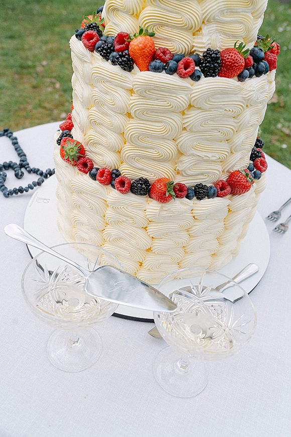 Wedding cake with piped buttercream frosting topped with fresh berries on a stand beside coupe glasses and knife on a lawn table