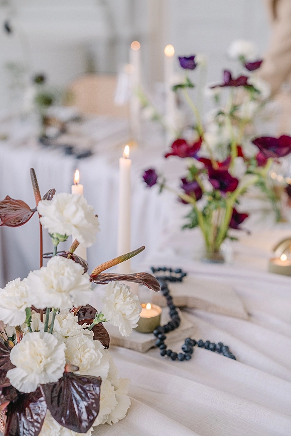 Reception tablescape with wedding table centerpiece of white carnations, purple blooms, anthurium leaves, and candlelight on white linen in soft light