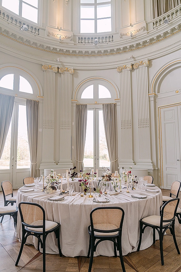 Reception tablescape with white linen, bud vase florals, and taper candles on a round table in a grand ballroom with arched windows