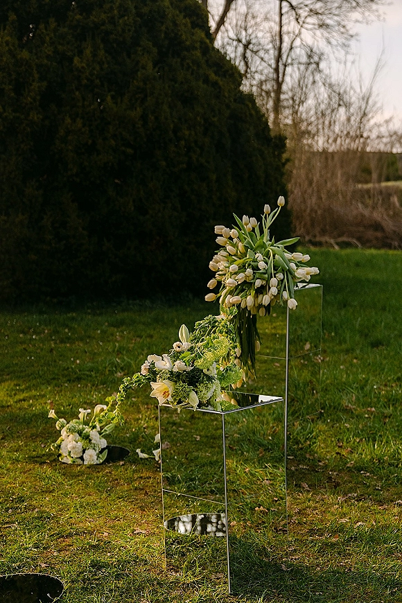 Ceremony floral decor with wedding ceremony flowers in white tulips on a clear acrylic pedestal, set on a grass lawn by trees under open sky