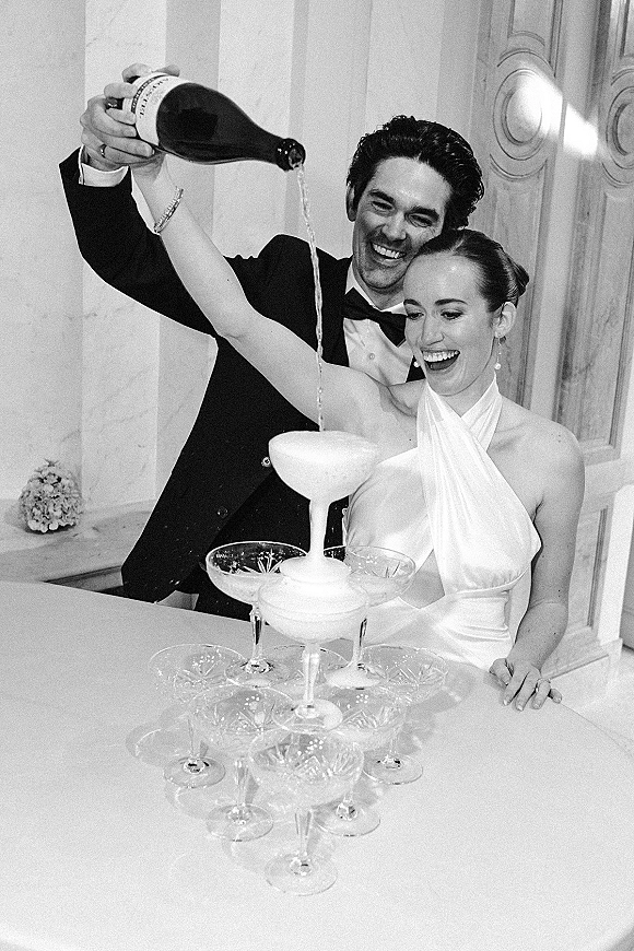 Champagne tower moment as bride in halter wedding dress pours from a bottle beside groom in tuxedo by a reception bar indoors