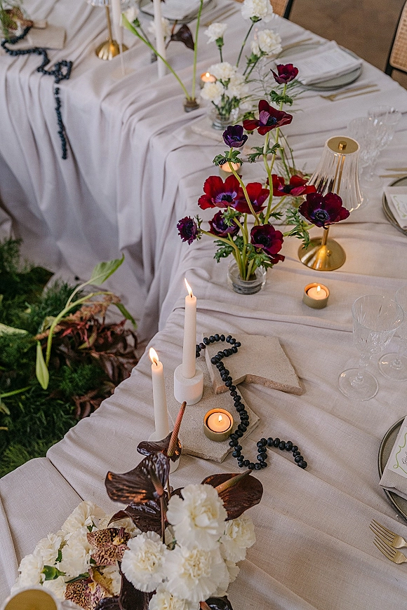 Reception tablescape with wedding table decor featuring linen, red anemones, taper and tealight candles, gold flatware, and greenery on grass