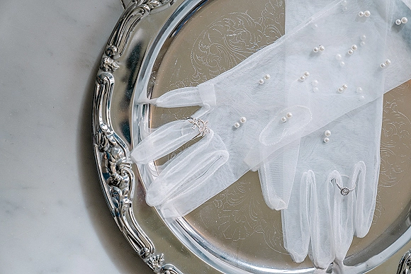 Bridal gloves in white tulle with pearl embellishments, styled with an engagement ring on a silver tray atop a light gray surface