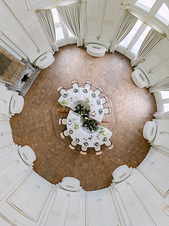 Reception table setup with a round wedding reception table, white linens, place settings, and floral centerpieces near a fireplace and arched windows