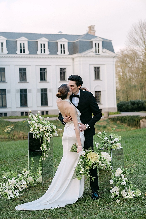 Wedding kiss portrait of bride and groom kissing on a garden lawn, bride in strapless satin gown holding white tulip bouquet before a manor house