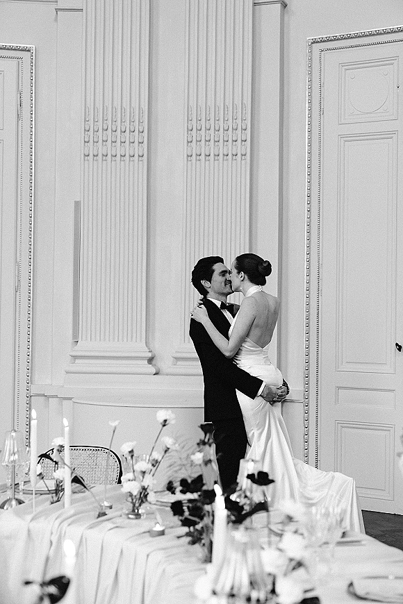 Wedding kiss as groom lifts bride in a tuxedo beside a candlelit reception table, framed by ornate walls and decorative columns