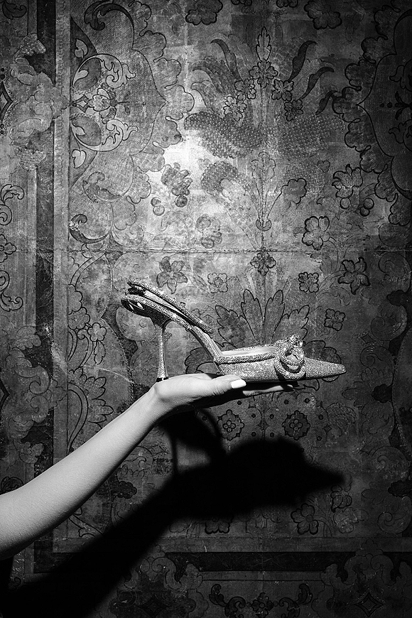 Bridal shoes sparkle as silver glitter high heel sandals with a rhinestone buckle are held in hand against patterned wallpaper