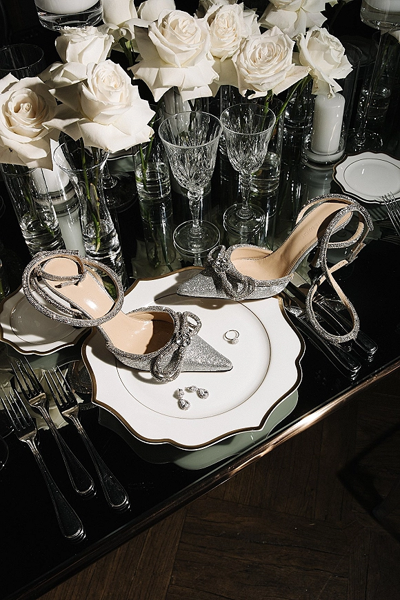 Bridal shoes, silver glitter wedding shoes with rhinestone bow on a mirrored tabletop beside engagement ring, roses, and candles
