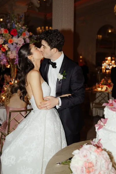 Wedding kiss as bride in strapless lace gown and groom in tux lean in by white cake with pink sugar flowers in a grand ballroom reception