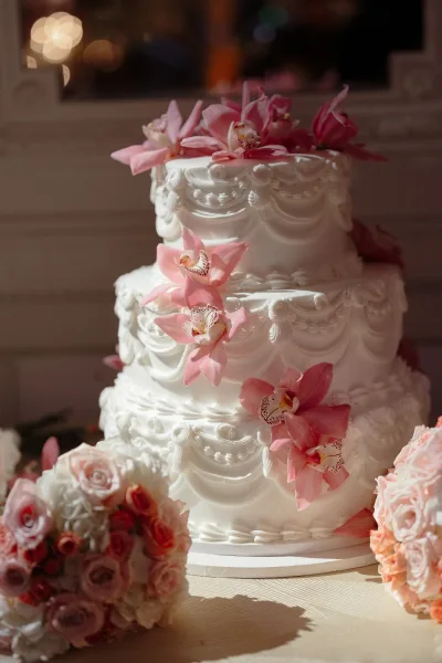 Wedding cake three tier wedding cake with ornate white buttercream piping and pink orchid accent on a stand by window light on a wooden table