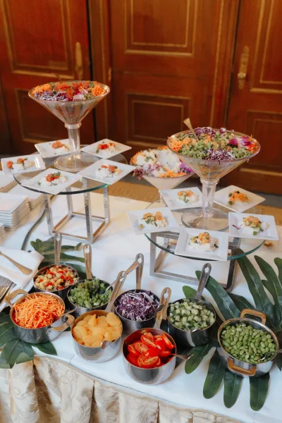 Wedding food display with a wedding appetizer station on glass risers, martini glass bowls and square plates over a leafy runner indoors