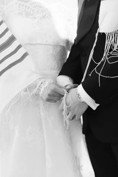 Wedding ceremony moment with bride and groom holding hands, ring and wristwatch visible, lace sleeves and tallit fringes in bright light