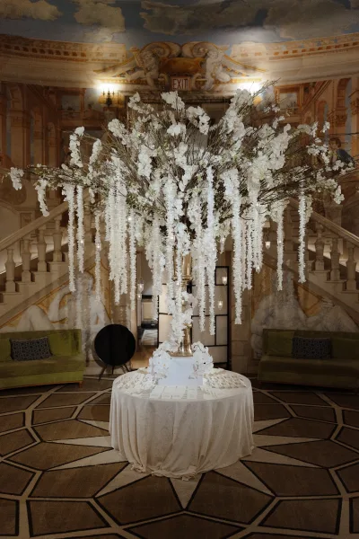 Wedding escort card table with tall floral centerpiece in a gold urn, hanging white florals and votive candles before a grand staircase