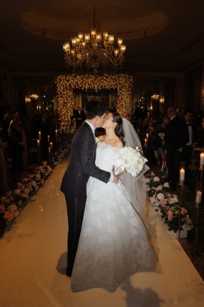 Wedding kiss as bride in lace dress and veil holds a white bouquet, groom in tuxedo beneath a floral arch in a candlelit ballroom