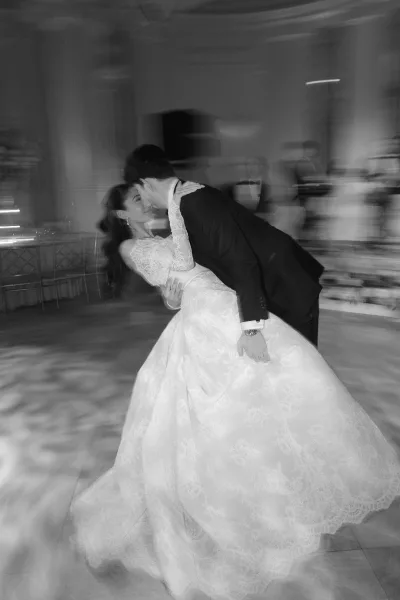 First dance kiss as the groom dips the bride in a lace long-sleeve dress and veil on a ballroom dance floor, guests blurred in lights