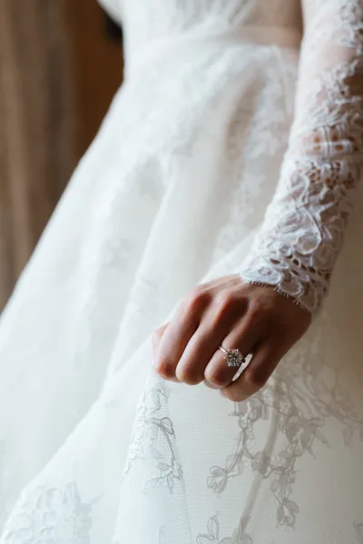 Engagement ring with a diamond solitaire ring on a hand resting on a long sleeve lace wedding dress in soft indoor light