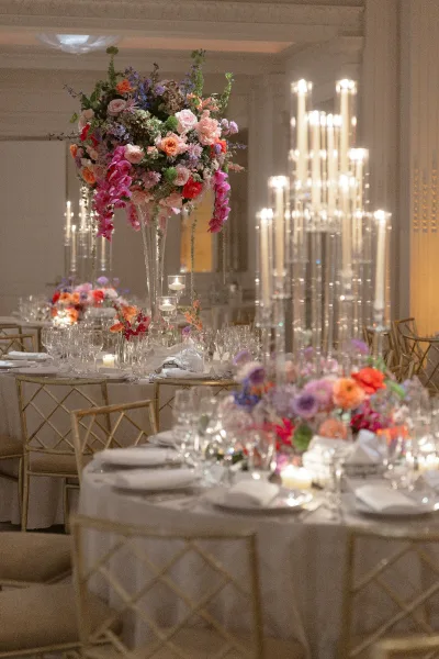 Reception tablescape with tall wedding centerpieces, crystal candelabra and taper candles on round linens in a warm-lit ballroom with gold chairs
