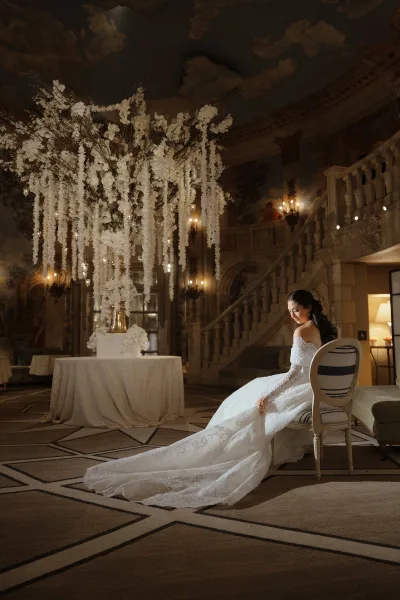 Bridal portrait of a bride in a lace wedding dress with an off-shoulder neckline and long train, seated in an ornate ballroom by a grand staircase.
