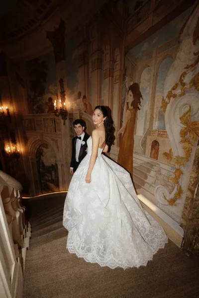 Wedding couple portrait with bride on staircase in a strapless lace ball gown, groom behind her in tuxedo under warm sconces and mural wall