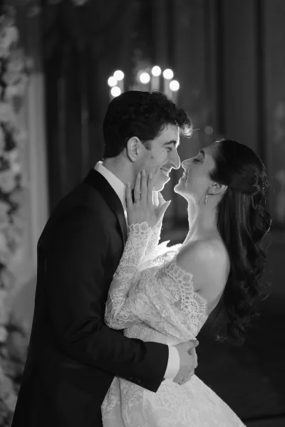 Wedding couple portrait in black and white, bride with lace sleeves touches groom’s face, showing ring beneath chandeliers and columns indoors
