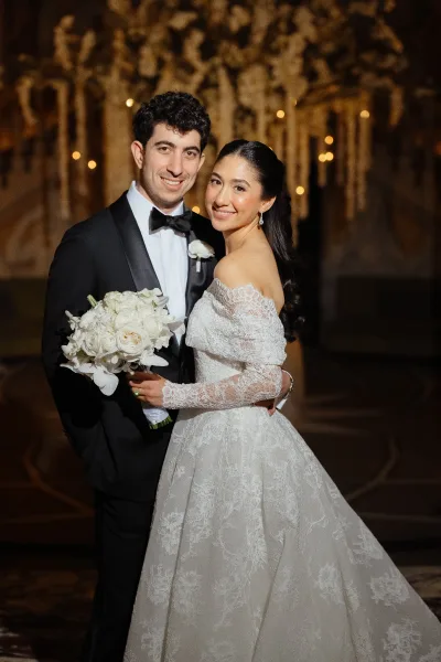Couple portrait of bride and groom arm in arm, bride holding white rose and orchid bouquet under warm bistro lights outdoors at night