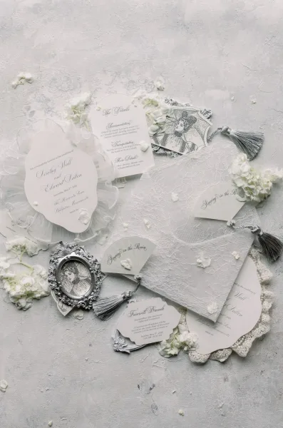 Invitation suite wedding invitation flat lay with oval cards, lace, tassels, white flowers, and silver frames on a light gray textured surface