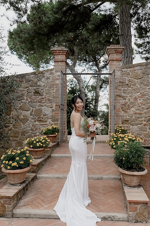 Bridal portrait of a bride holding bouquet, looking over her shoulder in a strapless satin gown beside a stone wall and iron gate