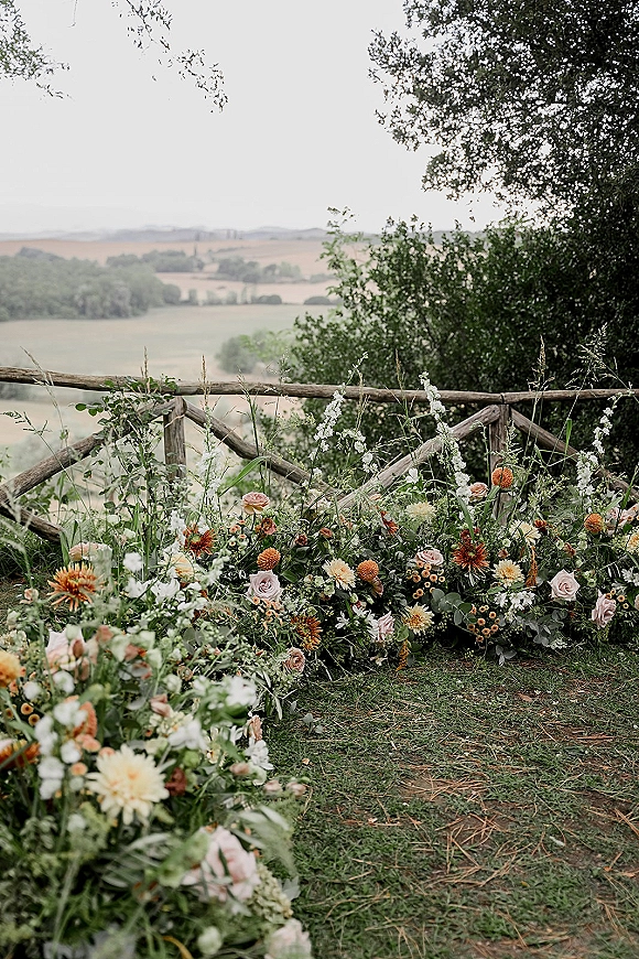 Ceremony aisle flowers border a grass path with roses, dahlias, and wildflower greenery, framed by a rustic wooden fence and rolling hills