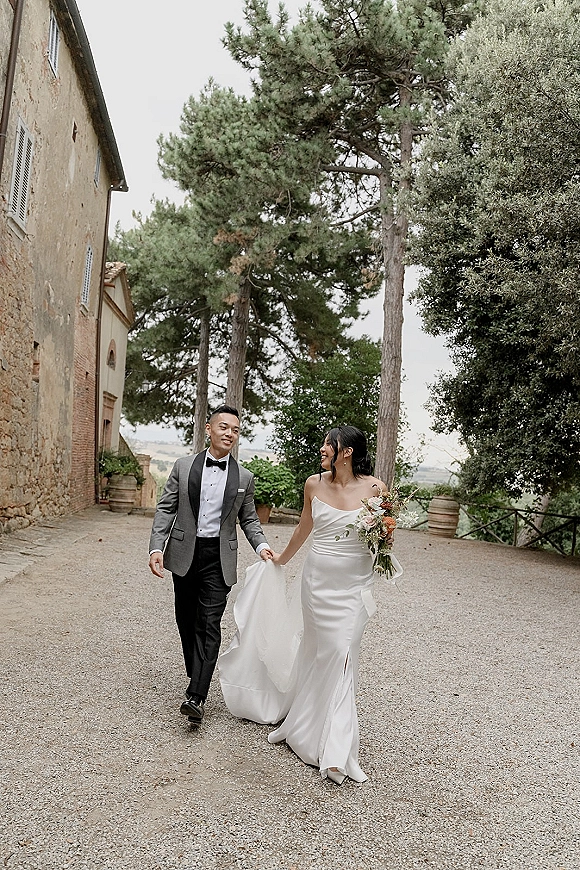 Couple portrait of bride and groom walking hand in hand, bride holding bouquet on a gravel path by a stone building under trees