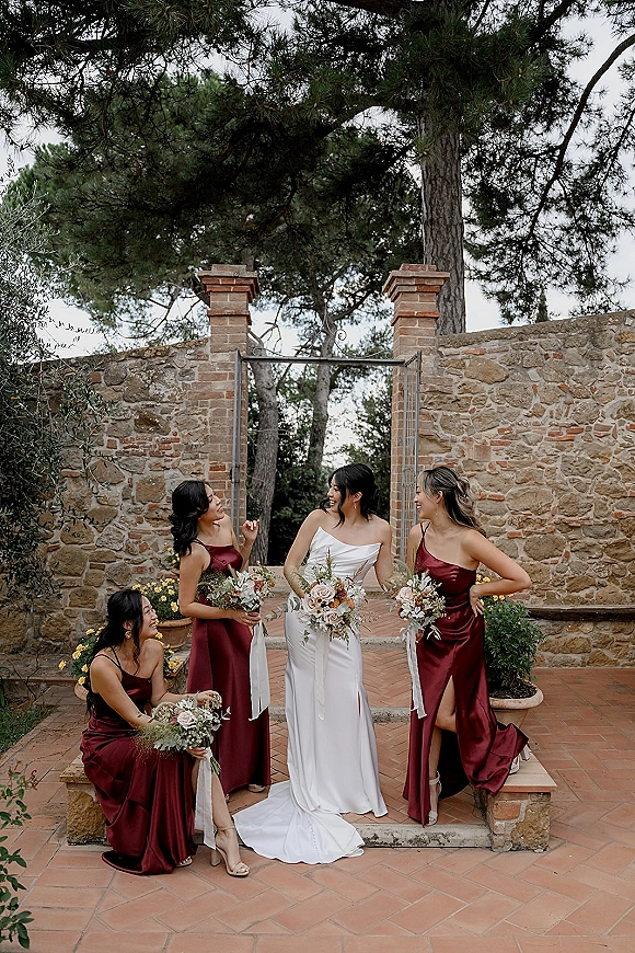 Bride with bridesmaids laughing in burgundy dresses, holding bouquets with ribbon streamers on stone steps by an iron gate courtyard