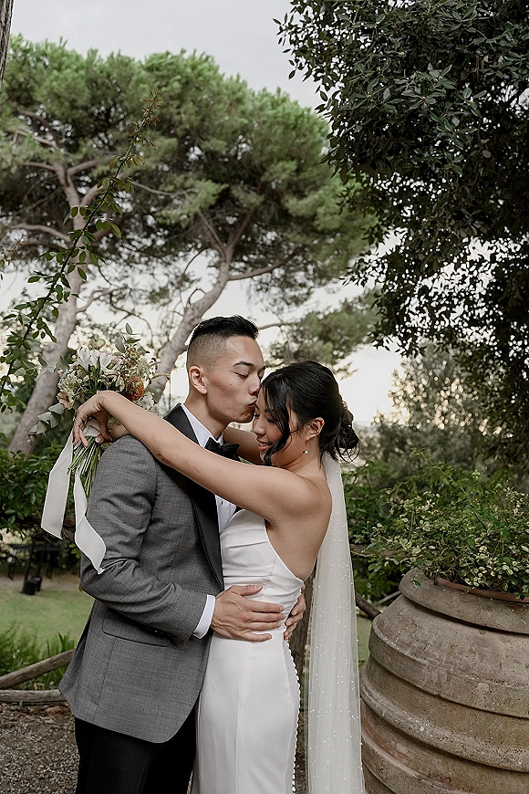 Couple portrait of bride and groom in an embrace, bride holding a ribbon-tied bouquet and veil as he kisses her forehead in a garden setting