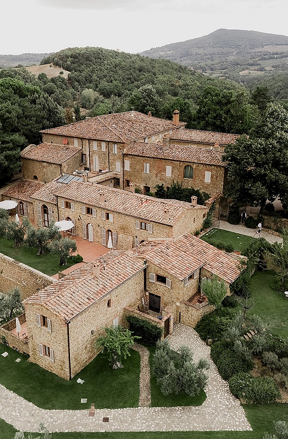 Wedding venue exterior, a stone villa with terracotta roof and arched doors, patio umbrellas and olive trees, set against forested hills