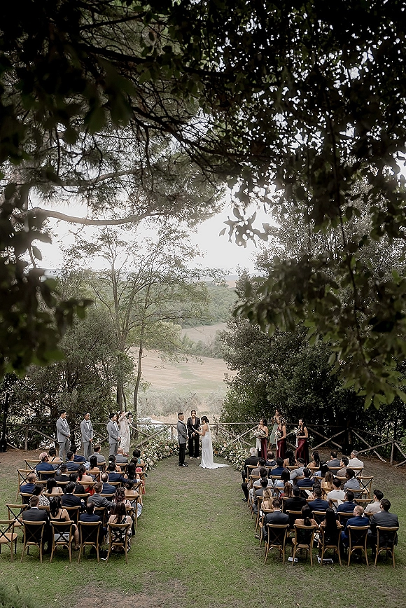 Outdoor wedding ceremony with garden ceremony setup, bride and groom at the altar beside floral aisle decor under a tree canopy