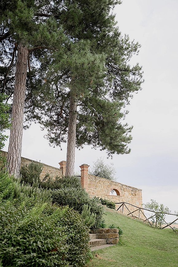 Outdoor wedding venue with stone wall accent, brick pillars and stone steps, framed by pine trees, shrubs, and an overcast sky
