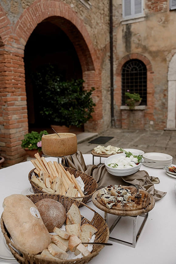 Wedding cocktail hour appetizer table with bread baskets and a cheese wheel on a linen runner in a stone courtyard under a brick archway.