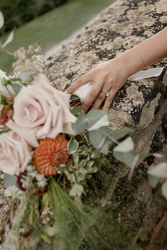 Engagement ring sparkling on a hand beside a diamond engagement ring bracelet, nestled against a blush rose and eucalyptus bouquet on a stone ledge