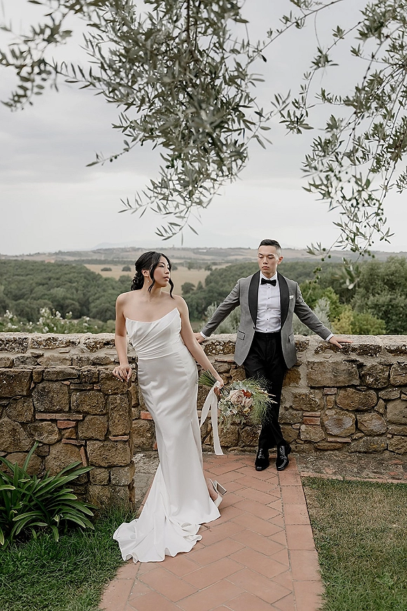 Couple portrait of bride in strapless satin wedding dress holding a bouquet with trailing ribbons, groom in tuxedo by a stone wall courtyard