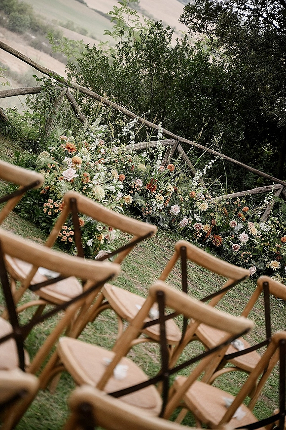 Ceremony aisle flowers with grounded ceremony florals of roses, wildflowers, and greenery lining wooden chairs on a grassy hillside lawn