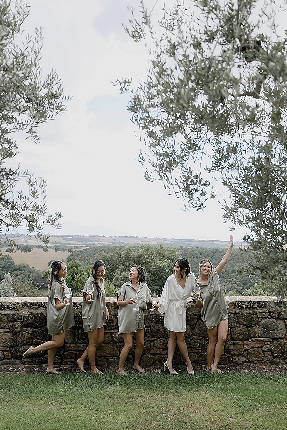 Bridesmaid group photo with bridesmaids getting ready in robes, holding champagne flutes beside a stone wall with olive trees and hills