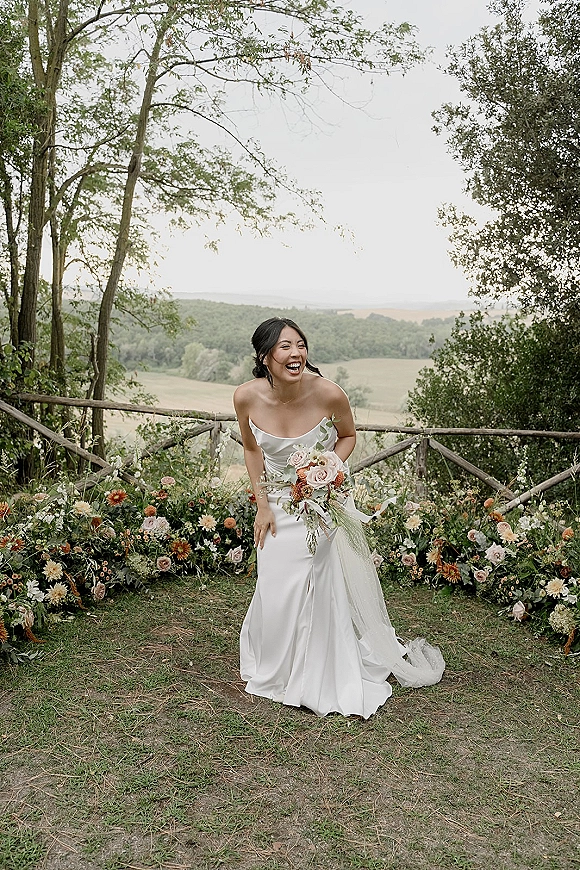 Bridal portrait of a laughing bride holding a bouquet in a strapless satin gown, with ribbon and wildflower ground blooms by a wooden fence and hillside field