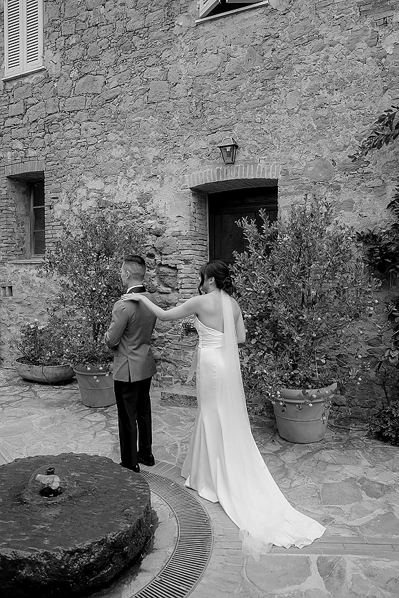 Wedding first look as the bride taps the groom’s shoulder, both seen from behind in a stone courtyard with potted plants and a wooden door