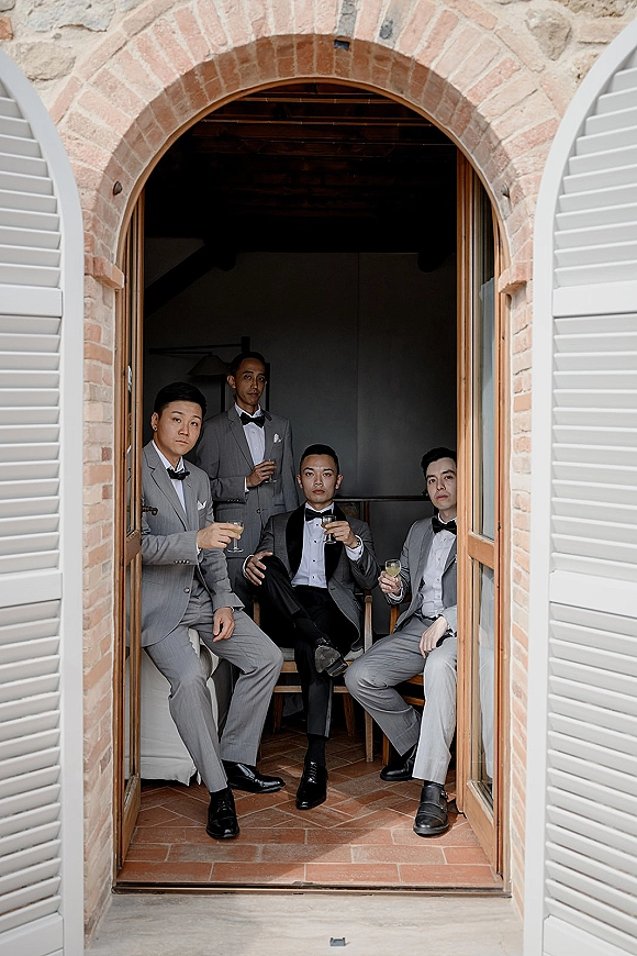 Groomsmen portrait with groomsmen in gray suits holding champagne coupes, posed on wooden chairs by a brick arch doorway indoors
