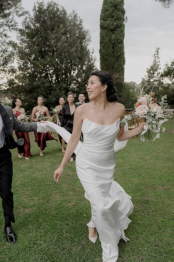 Wedding party portrait of bride and groom walking on an outdoor lawn, bride holding a pastel bouquet with bridesmaids in burgundy dresses behind