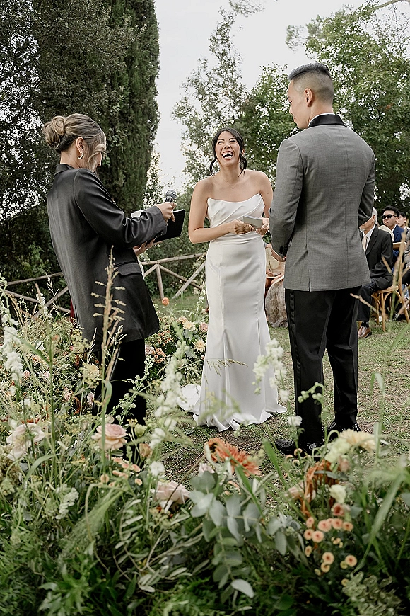 Wedding vows during an outdoor wedding ceremony as the bride reads from a vow book beside the groom, with low florals and guests on a garden lawn